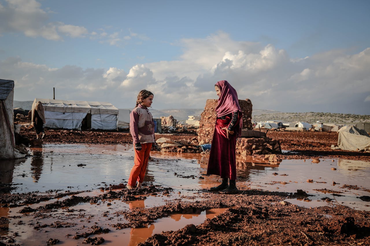 Mastering the First Impression: Your intriguing post title goes here Two children talk beside puddles in an Idlib refugee camp, surrounded by tents and muddy terrain.